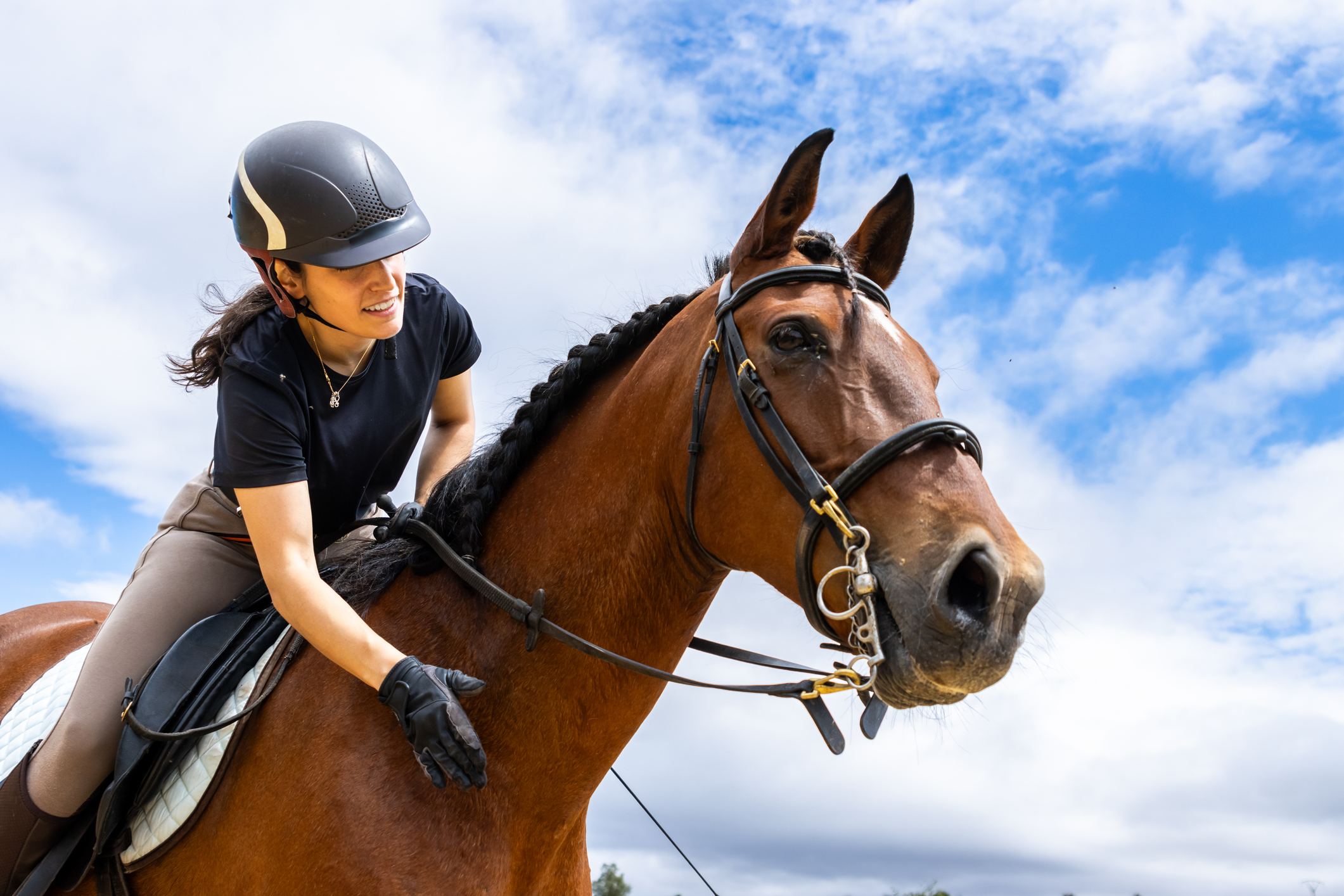 Young woman wearing a helmet and riding gloves is riding a horse and petting it under a cloudy sky