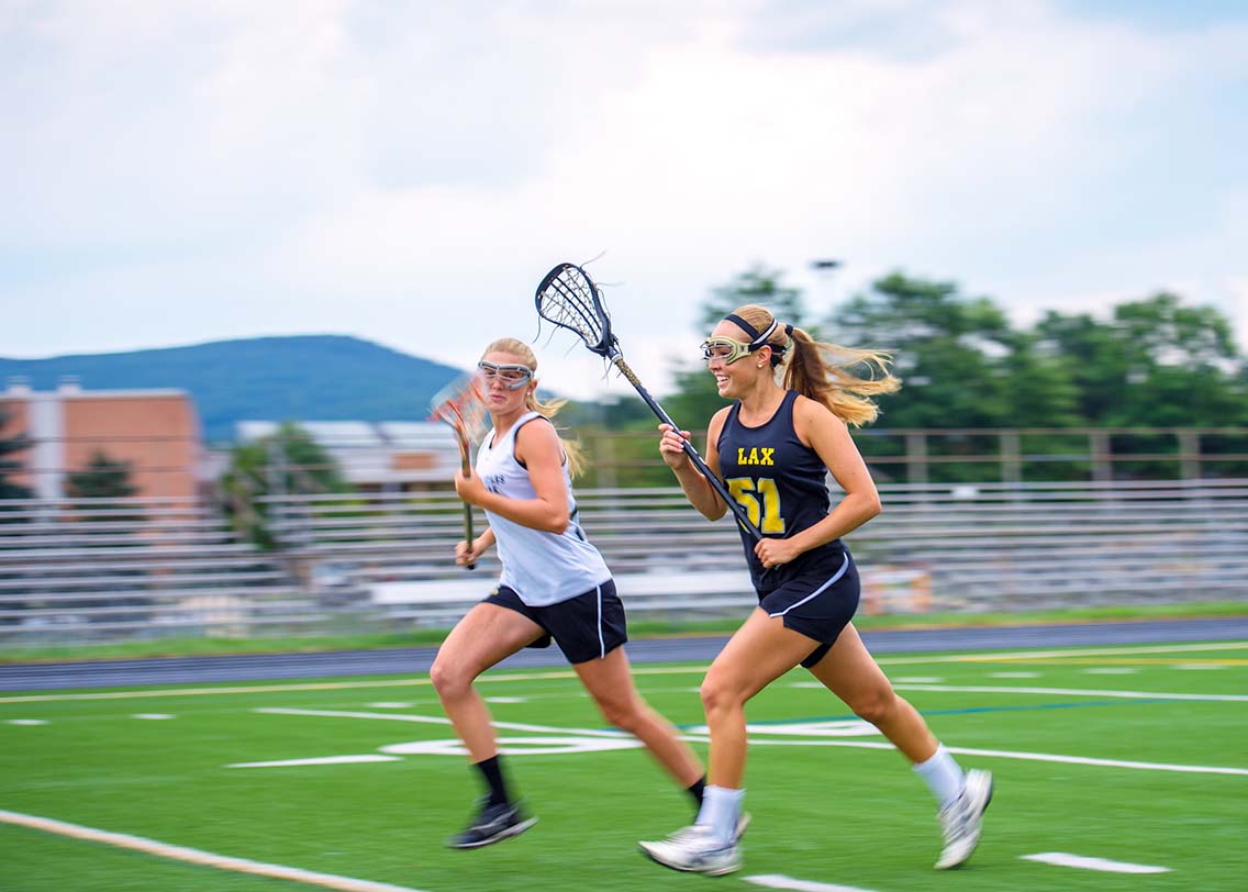 2 women running with lacrosse sticks on pitch