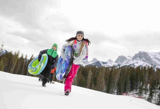 Happy brother and sister running with inner tubes below snowy mountain